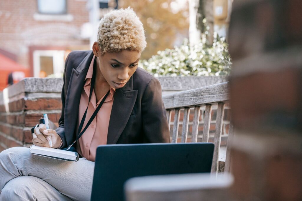 Woman sitting on a bench with a notepad looking down at a laptop.
