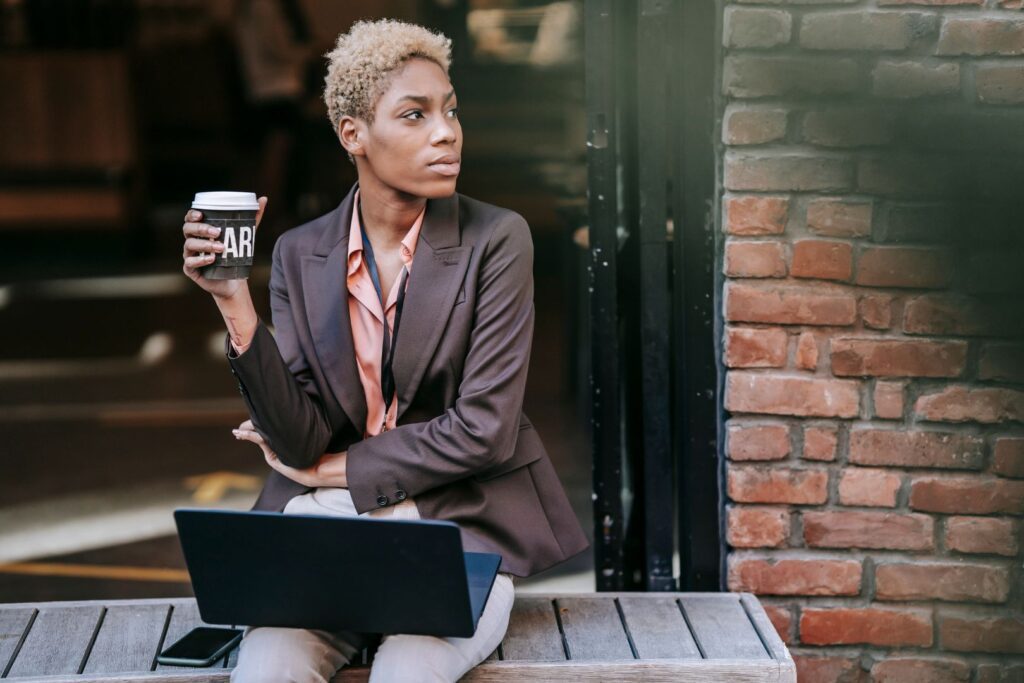 Woman sitting with coffee and a laptop Are blogs still relevant for your business in 2023? businesswoman holding a takeaway cup of coffee and looking into the distance with a laptop on her knee and mobile phone on a bench beside her.